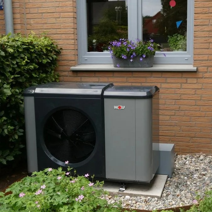 Eine graue Wärmepumpe steht vor einem Fenster mit lila Blumen und grünem Busch