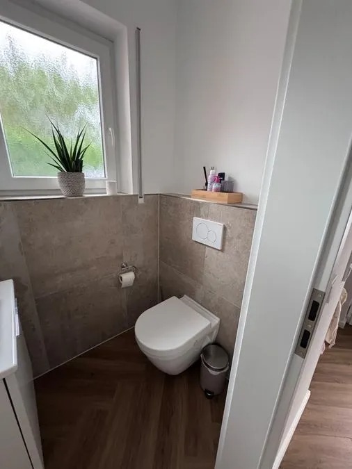 Modern weißes Bad mit Wandtoilette, Holzboden, Pflanze und Kosmetikartikeln am Fenster