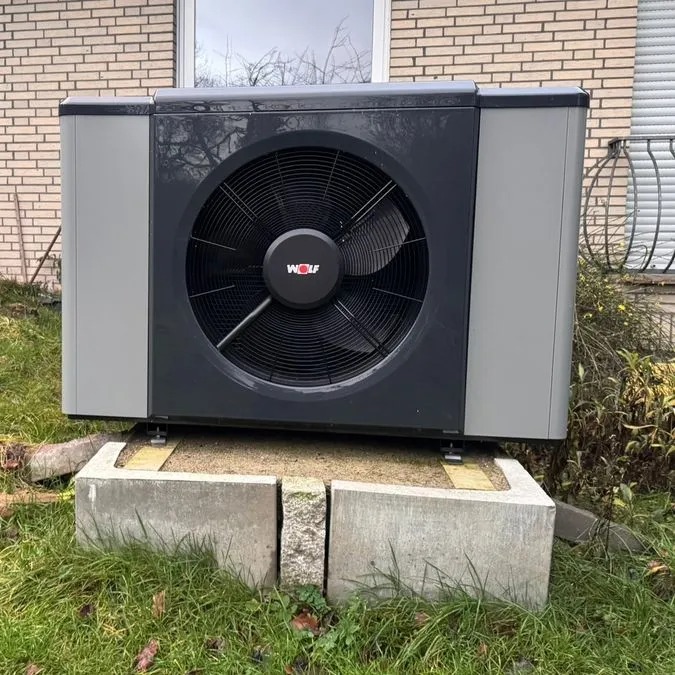 Eine große grauschwarze Wärmepumpe von Wolf steht auf Betonblöcken vor einem Haus