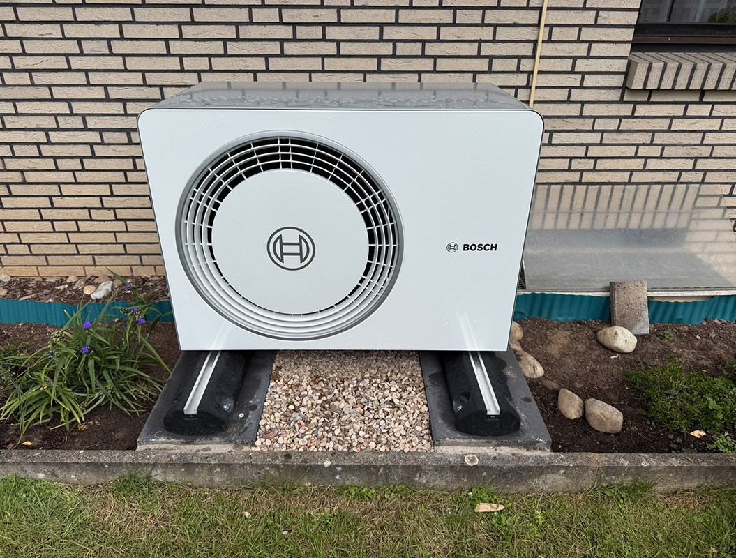 Eine weißgraue Bosch-Wärmepumpe steht vor einer Backsteinwand im Garten