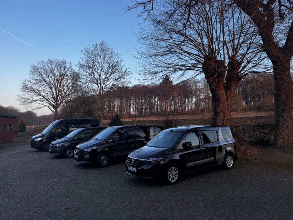 Vier schwarze Fahrzeuge mit Wessels Logo stehen nebeneinander auf einem Parkplatz unter kahlen Bäumen